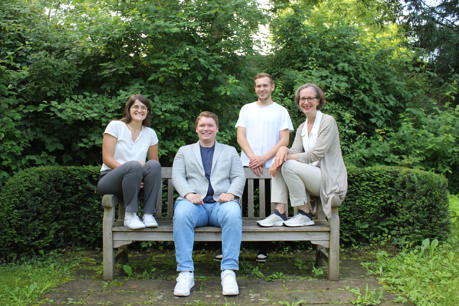 Auf dem Bild sind vier Menschen zu sehen die auf und an einer Bank sitzen und stehen. Es sind zwei Frauen und zwei Männer.