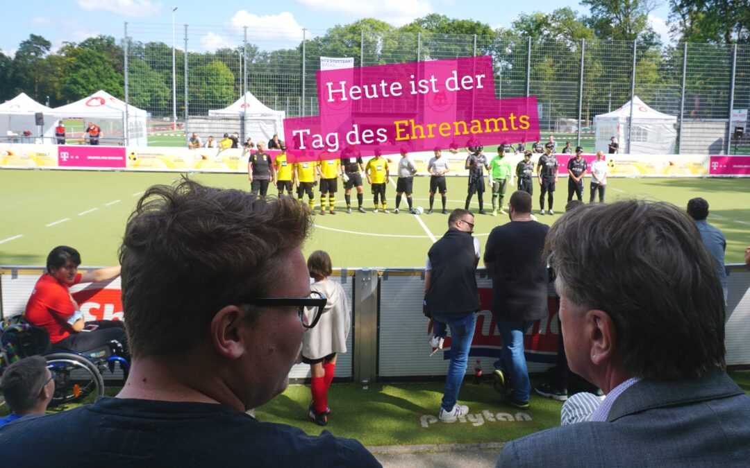 Landtagsabgeordneter Felix Herkens mit Sozialminister Manne Lucha von hinten. Sie blicken auf ein Fußballfeld. Spieler der Blindenfußball Bundesliga sind in einer Reihe auf dem Spielfeld aufgestellt.